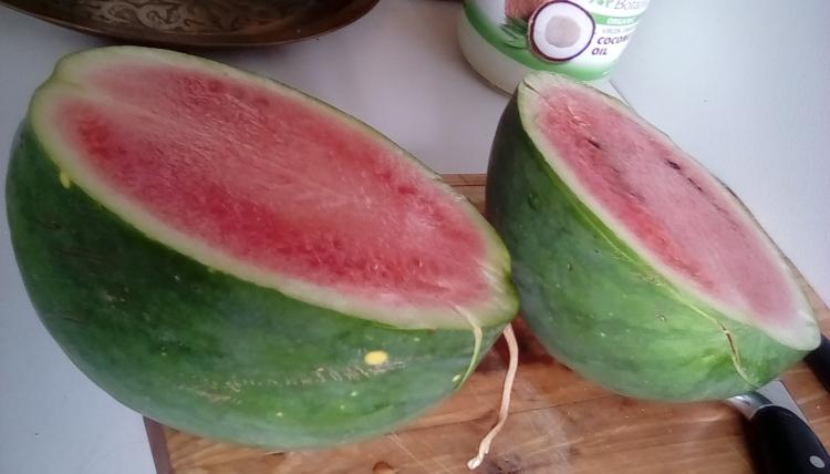 Weeks NC Giant cross watermelon fruit with stars, sliced in two, on a cutting board. It has pink flesh and dark seeds. The rind has stripes.